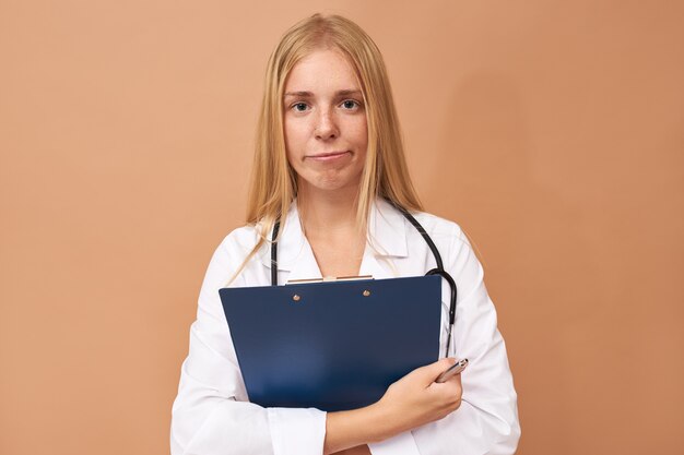 Beautiful experienced young female practitioner in white surgical coat holding clipboard