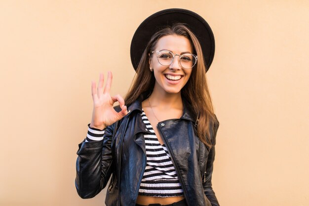 Beautiful fashion girl in leather jacket and black hat shows ok sign isolated on light yellow wall