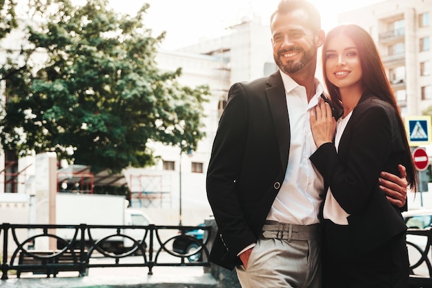 Free photo beautiful fashion woman and her handsome boyfriend in suit sexy smiling brunette model in black evening dress fashionable couple posing in the street at sunset brutal man and his female outdoors