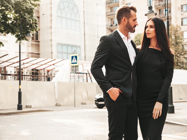 Beautiful fashion woman and her handsome elegant boyfriend in suit Sexy brunette model in black evening dress Fashionable couple posing in the street in Europe Brutal man and his female outdoors