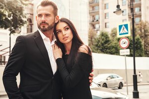 Beautiful fashion woman and her handsome elegant boyfriend in suit sexy brunette model in black evening dress fashionable couple posing in the street in europe brutal man and his female outdoors