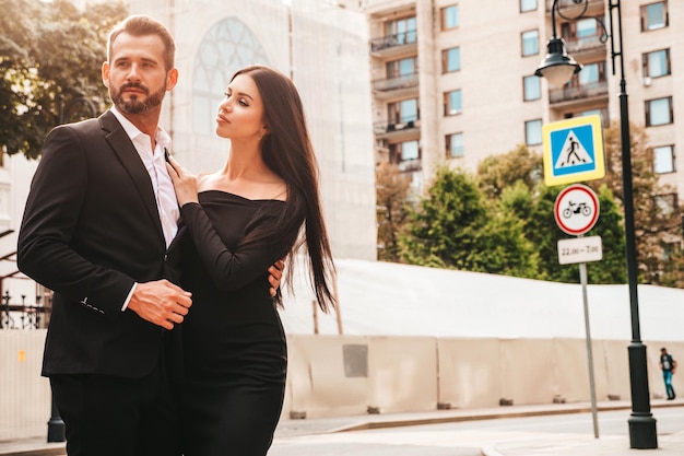 Bella donna alla moda e il suo fidanzato bello ed elegante in tuta modello bruna sexy in abito da sera nero coppia alla moda in posa per strada in europa uomo brutale e la sua femmina all'aperto