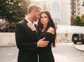 Beautiful fashion woman and her handsome elegant boyfriend in suit sexy brunette model in black evening dress fashionable couple posing in the street in europe brutal man and his female outdoors