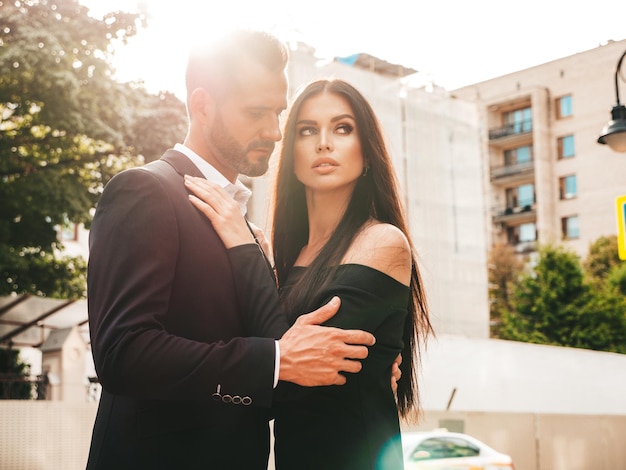 Beautiful fashion woman and her handsome elegant boyfriend in suit Sexy brunette model in black evening dress Fashionable couple posing in the street in Europe Brutal man and his female outdoors
