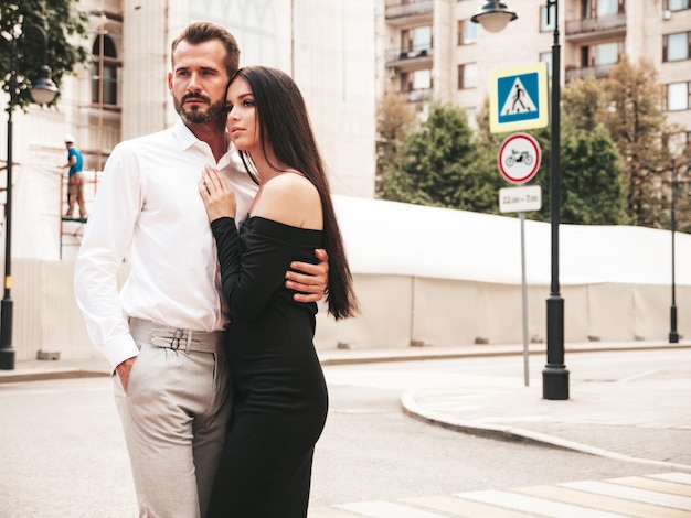 Beautiful fashion woman and her handsome elegant boyfriend in suit Sexy brunette model in black evening dress Fashionable couple posing in the street in Europe Brutal man and his female outdoors
