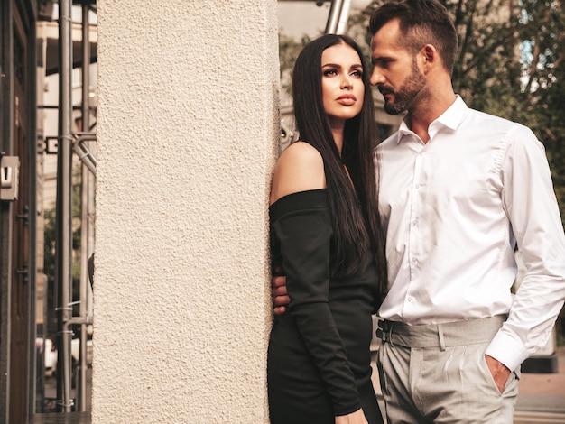 Free photo beautiful fashion woman and her handsome elegant boyfriend in suit sexy brunette model in black evening dress fashionable couple posing in the street in europe brutal man and his female outdoors