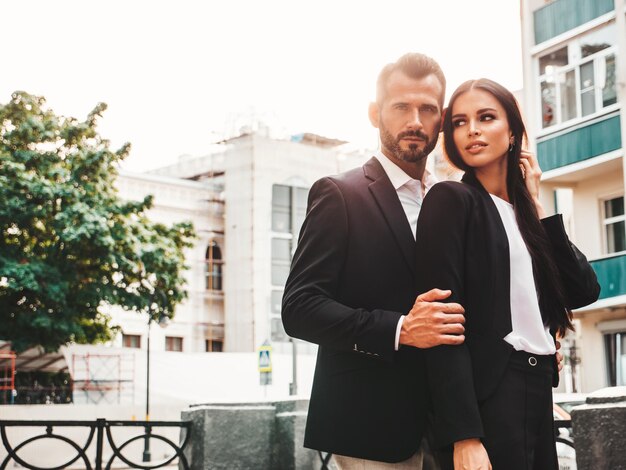 Beautiful fashion woman and her handsome elegant boyfriend in suit Sexy brunette model in black evening dress Fashionable couple posing in the street in Europe Brutal man and his female outdoors