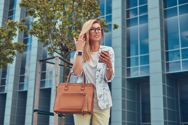 Красивая модная женщина-блогер в стильной одежде и очках с сумочкой держит смартфон, стоя против небоскреба.