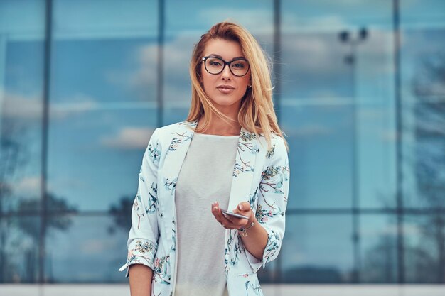 Красивая модная женщина-блогер в стильной одежде и очках с сумочкой держит смартфон, стоя против небоскреба.