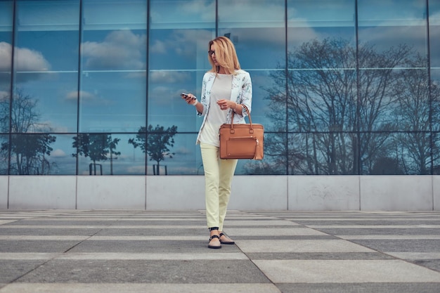 Красивая модная женщина-блогер в стильной одежде и очках с сумочкой, с помощью смартфона, стоящая на фоне небоскреба.