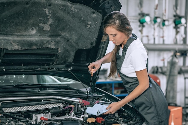 A beautiful female in a black jumpsuit and a white t-shirt, checking the oil level in a black car in the garage.
