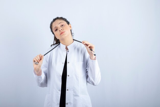 Beautiful female doctor in white coat posing with stethoscope over white wall. 