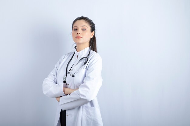 Beautiful female doctor in white coat standing arms crossed over white wall.