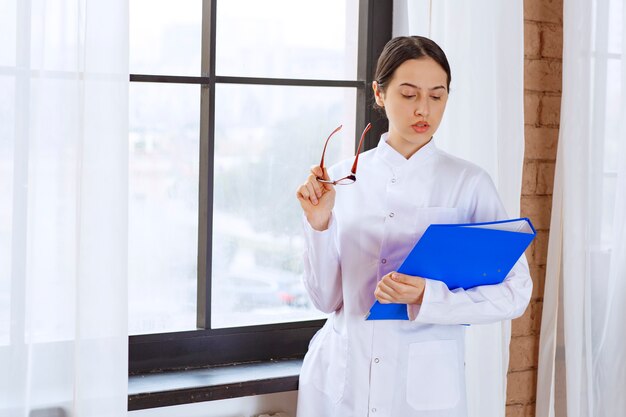 Beautiful female doctor in white coat with folder standing near the window. 