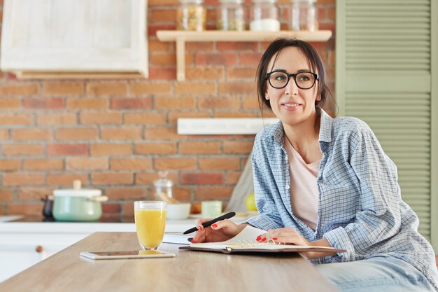 Beautiful female dressed casually, wears spectacles, sits at wooden kitchen desk,