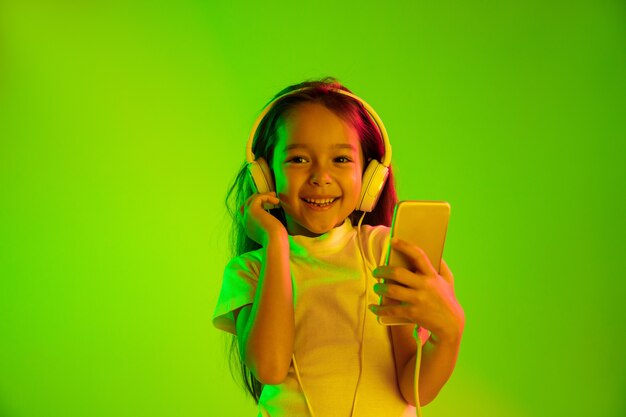 Beautiful female half-length portrait isolated on green backgroud in neon light. Young emotional girl. Human emotions, facial expression concept. Using smartphone for vlog, selfie, chating, gaming.