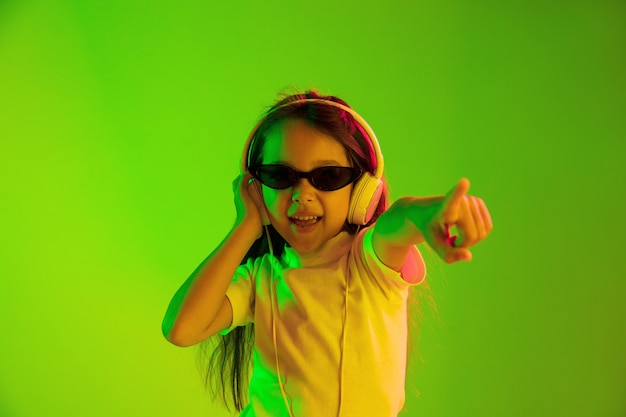 Free photo beautiful female half-length portrait isolated on green backgroud in neon light. young emotional teen girl. human emotions, facial expression concept. dancing in sunglasses and pointing up.