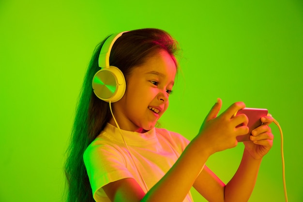 Красивый женский поясной портрет изолированный на зеленой стене в неоновом свете. Молодая эмоциональная девушка. Человеческие эмоции, концепция выражения лица. Использование смартфона для видеоблога, селфи, чата, игр.