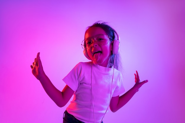 Beautiful female half-length portrait isolated on purple backgroud in neon light. Young emotional teen girl in eyeglasses. Human emotions, facial expression concept. Trendy colors. Dancing, smiling.