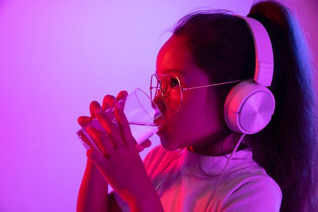 Beautiful female half-length portrait isolated on purple backgroud in neon light. Young emotional teen girl in eyeglasses. Human emotions, healthcare, facial expression concept. Drinking pure water.