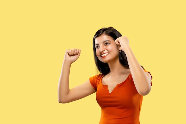 Beautiful female half-length portrait isolated on yellow  space. Young emotional indian woman in dress celebrating a win. Negative space