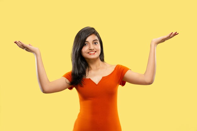 Beautiful female half-length portrait isolated on yellow  space. Young emotional indian woman in dress pointing and showing. Negative space