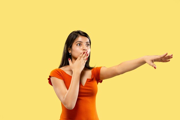 Beautiful female half-length portrait isolated on yellow studio background. Young emotional indian woman in dress pointing and showing. Negative space. Facial expression, human emotions concept.