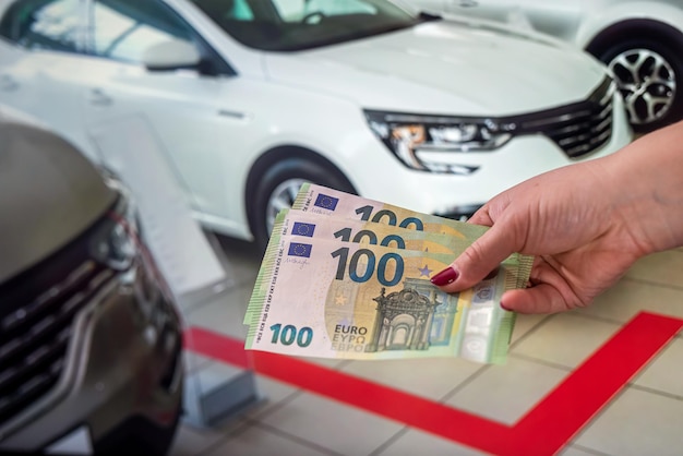 Красивая женская рука с ухоженными красными ногтями держит большое количество банкнот евро возле машины. концепция покупки и продажи автомобиля. концепция салона автомобиля