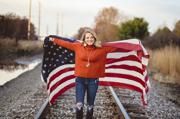 Bella femmina che tiene la bandiera americana mentre levandosi in piedi sulla ferrovia