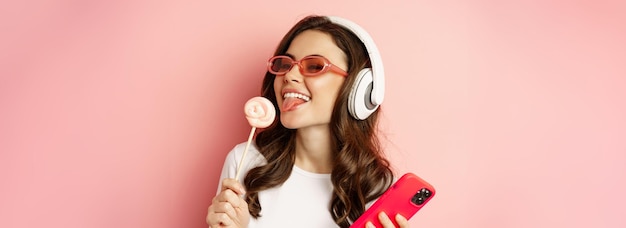 Бесплатное фото Красивая женская модель слушает музыку в наушниках, держа в руках леденец и мобильный телефон, позируя в песне