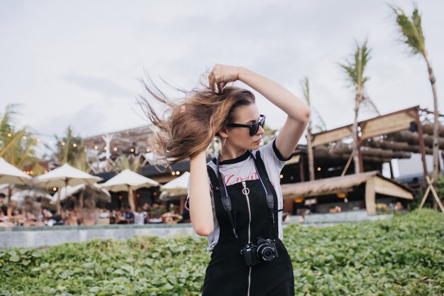 Beautiful female photographer in sunglasses playing with her hair. Outdoor shot of cheerful european girl walking in park with camera.