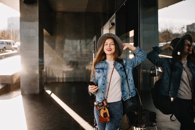 Beautiful female photographer in trendy outfit with black phone posing smiling near shopping center.