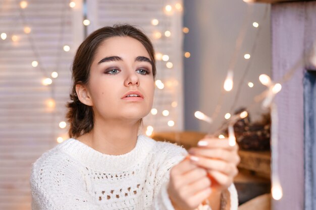 Beautiful female portrait of a young girl in a white sweater on a of garlands and bokeh.