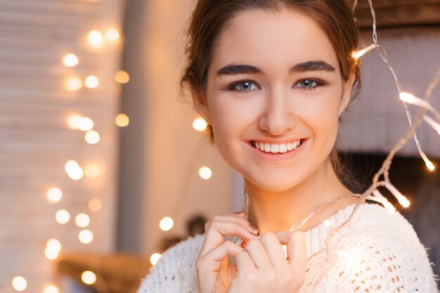 Beautiful female portrait of a young girl in a white sweater on a of garlands and bokeh.