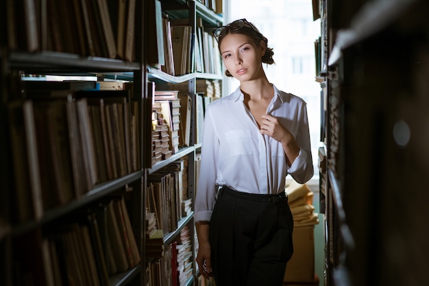 Красивая студентка в белой рубашке стоит между рядами в библиотеке