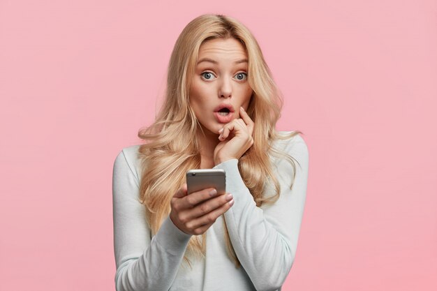 Beautiful female with blonde hair feels surprised as checks her email box on cell phone, wonders big discounts in web store, isolated over pink wall. Emotions and unexpectedness concept