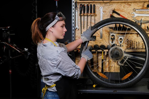 Beautiful female in working clothes, apron and goggles, repairs a bicycle wheel in a workshop.