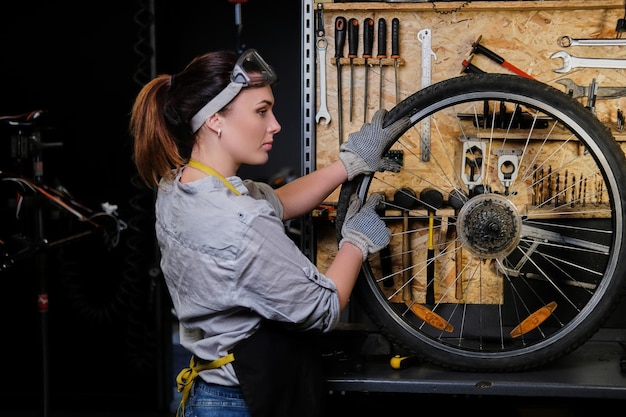 Beautiful female in working clothes, apron and goggles, repairs a bicycle wheel in a workshop.