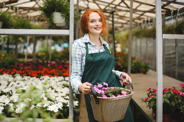 Beautiful florist in apron standing with metal basket with flowers in hands and happily looking in camera