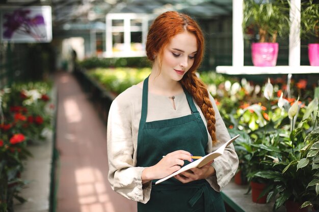 Beautiful florist in apron standing with pencil in hand and dreamily writing in notepad while working in greenhouse