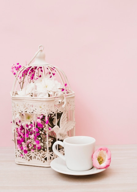 Красивые цветы в белой клетке с чашкой и блюдцем на деревянный стол с розовым фоном