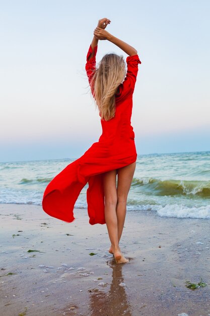 Красивая свободная женщина в красном платье на ветру на морском пляже гуляет летом
