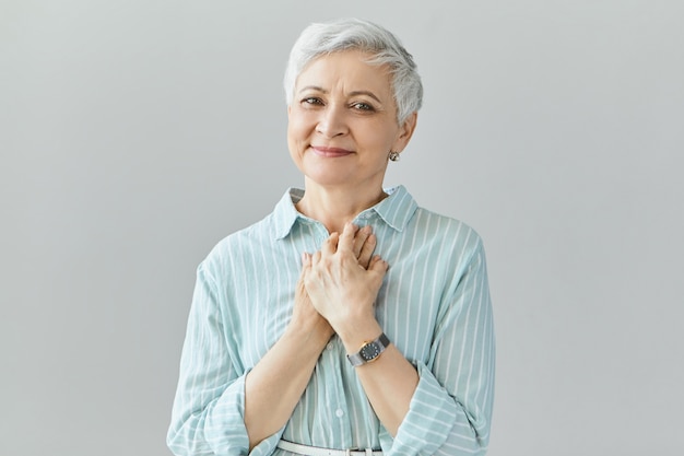 Beautiful friendly looking middle aged woman with sincere smile, expressing gratitude, feeling thankful, showing her heart filled with love, keeping hands on her chest. Positive genuine human feelings