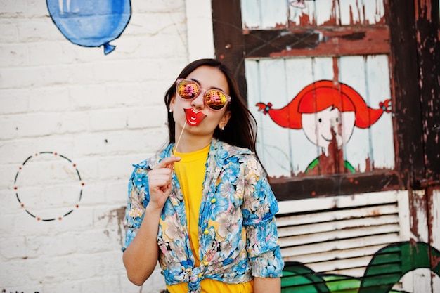 Free photo beautiful fun teenage girl with sunglasses and lips on stick wear yellow tshirt near graffiti wall