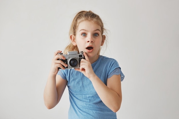 Beautiful funny little girl with blond hair and blue eyes holding photo camera in hands, looking straight with frightened expression.