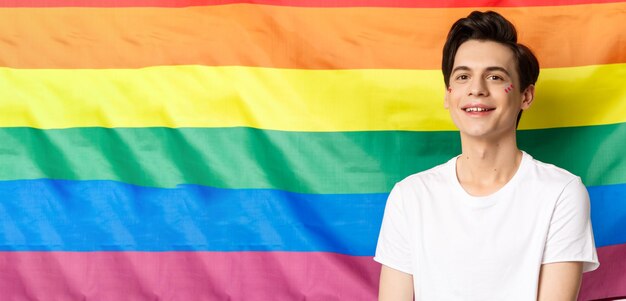 Beautiful gay man with glitter on face smiling happy and proud at camera standing against rainbow pr