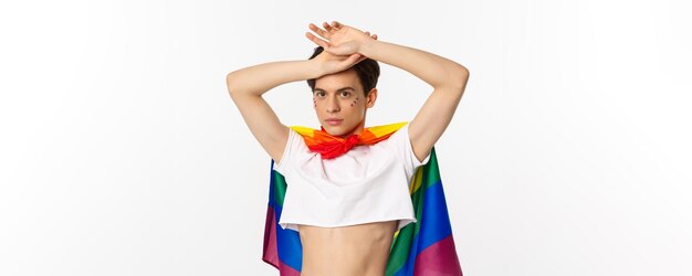 Beautiful gay man with glitter on face wearing crop top and rainbow lgbt flag posing against white b
