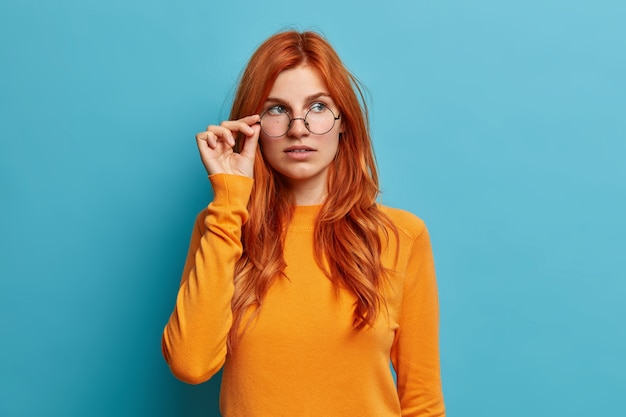 Beautiful ginger female student thinks about future project keeps hand on spectacles concentrated pensively aside dressed in casual orange jumper.