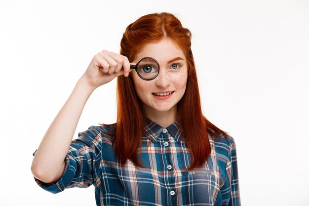 beautiful ginger girl with magnifier over white wall.