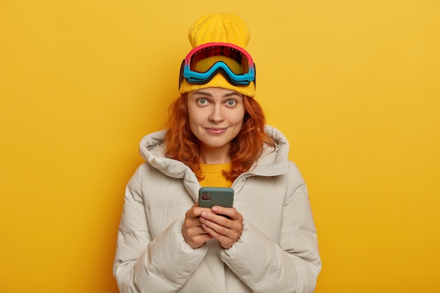 美しい生姜の女性は素晴らしい一日の後にソーシャルネットワークに写真を投稿し、冬の間アクティブな休息を取り、携帯電話を持ち、帽子、コート、保護スキーグラスを着用し、黄色い壁の上でポーズをとる